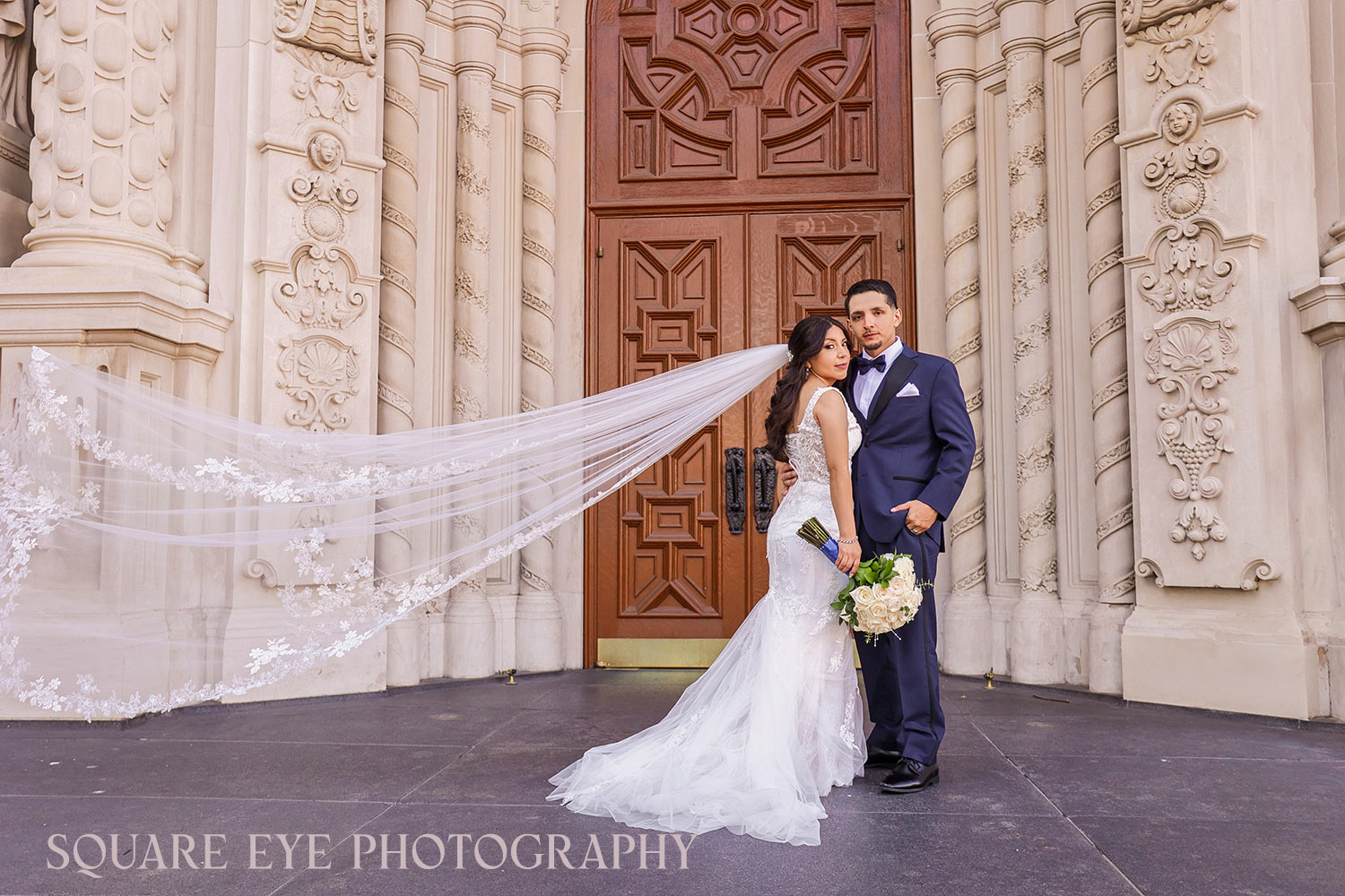 Couple-in-front-of-church-doors-with-veil-flying Cathedral veil wedding photos featuring a bride and groom with a dramatic flying veil in front of church doors, photographed by Square Eye Photography.